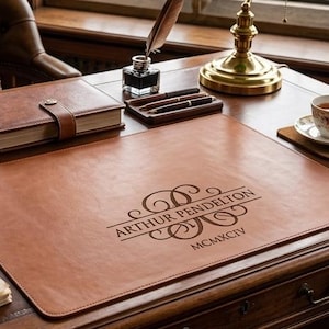 May include: A rectangular brown leather desk mat featuring a custom monogram with the name Arthur Pendelton and the Roman numeral MCMXCIV engraved in the center, placed on a wooden desk with books, a green banker lamp, and stationery.