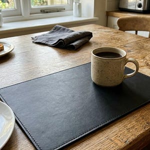 May include: A rectangular black leather placemat with stitched edges, featuring a speckled beige ceramic mug filled with dark liquid placed on its surface. The placemat is positioned on a wooden table next to a white plate with a piece of toast.