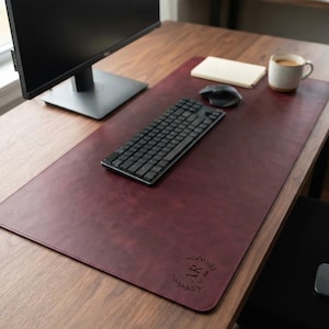 May include: A large, rectangular, dark burgundy desk mat with rounded corners. The mat features a debossed monogram of the letters AR surrounded by a laurel wreath design in the bottom right corner. The surface has a smooth, matte texture.