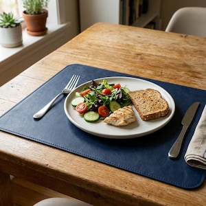 Könnte beinhalten: Eine rechteckige Platzmatte aus marineblauem Kunstleder mit abgerundeten Ecken und genähten Kanten. Auf einem weißen Keramikteller liegt ein Salat aus grünem Blattgemüse, Gurken und Kirschtomaten sowie gegrilltes Hähnchen und Toast.