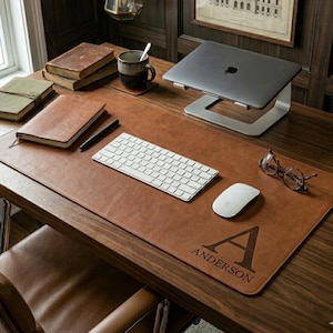 May include: A large rectangular brown leather desk mat featuring a personalized engraving of the letter A and the name Anderson. The mat is placed on a wooden desk with a white keyboard, a white computer mouse, and a pair of glasses.