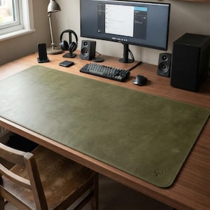 May include: A large, rectangular olive green desk mat made of smooth material with stitched edges and a small embossed tree logo in the bottom right corner, positioned on a wooden desk surface in front of a computer monitor and keyboard.