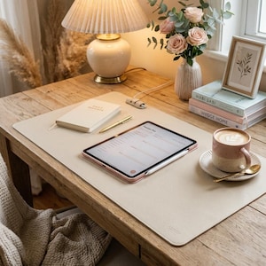 May include: A rectangular beige desk mat with a textured surface lies on a wooden table. On the mat are a white notebook, a gold pen, and a tablet displaying a planner. Nearby are a lamp, a vase with pink roses, and a coffee mug.