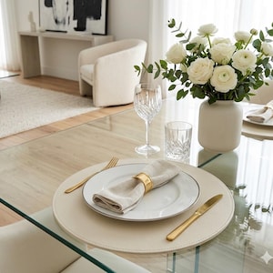 May include: A dining table setting with a white plate, gold cutlery, and a beige napkin held by a gold ring. A white vase holds white roses and greenery. A crystal wine glass and tumbler are also on the table.