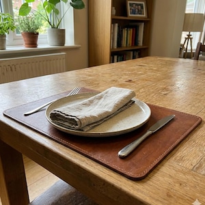May include: A rectangular brown leather placemat featuring stitched edges, topped with a speckled off-white ceramic plate, a folded beige linen napkin, a silver fork, and a silver knife, all arranged on a light-colored wooden dining table.