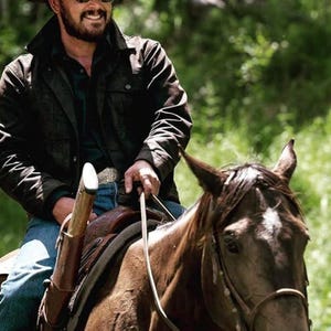 May include: A person wearing a black jacket, sunglasses, and a cowboy hat rides a brown horse. The horse has a white marking on its forehead. A rifle is mounted on the saddle. The background is blurred green foliage.