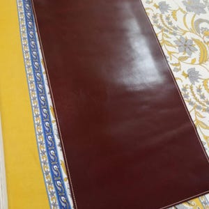 May include: A rectangular dark brown desk mat with rounded corners and white stitching along the entire perimeter. The surface is smooth and solid in color, featuring a clean, flat design with a consistent finish across the entire rectangular area.