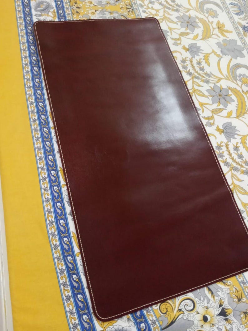 May include: A rectangular dark brown desk mat with rounded corners and white stitching along the entire perimeter. The surface is smooth and solid in color, featuring a clean, flat design with a consistent finish across the entire rectangular area.