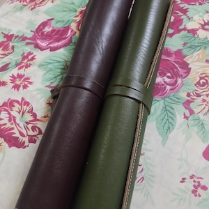 May include: Two cylindrical leather rolls, one in dark brown and one in olive green, each secured with a matching leather strap. The rolls feature visible stitching along the edges and a smooth, matte finish, presenting a structured, tubular design.