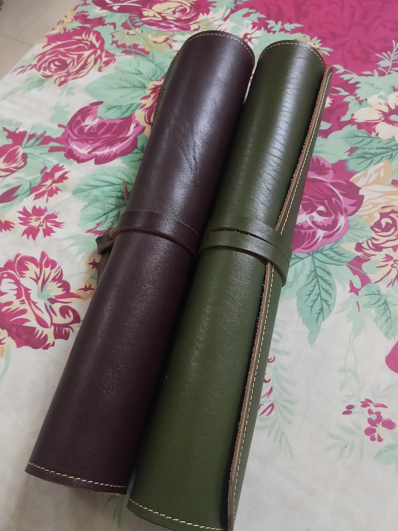 May include: Two cylindrical leather rolls, one in dark brown and one in olive green, each secured with a matching leather strap. The rolls feature visible stitching along the edges and a smooth, matte finish, presenting a structured, tubular design.