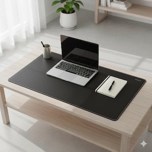 May include: A large rectangular black desk mat with rounded corners, featuring a silver laptop, a white notebook with a black pen, and a grey cylindrical pen holder placed on its surface. The mat has a smooth, matte finish.