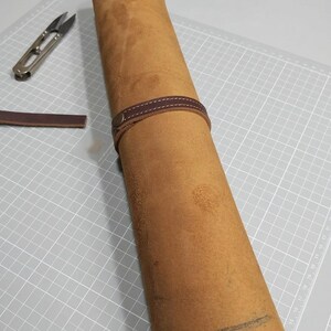 May include: A tan leather roll secured with a dark brown leather strap and a metal snap button. A small pair of metal snips and a loose strip of dark brown leather are positioned nearby on a white cutting mat with a grid pattern.