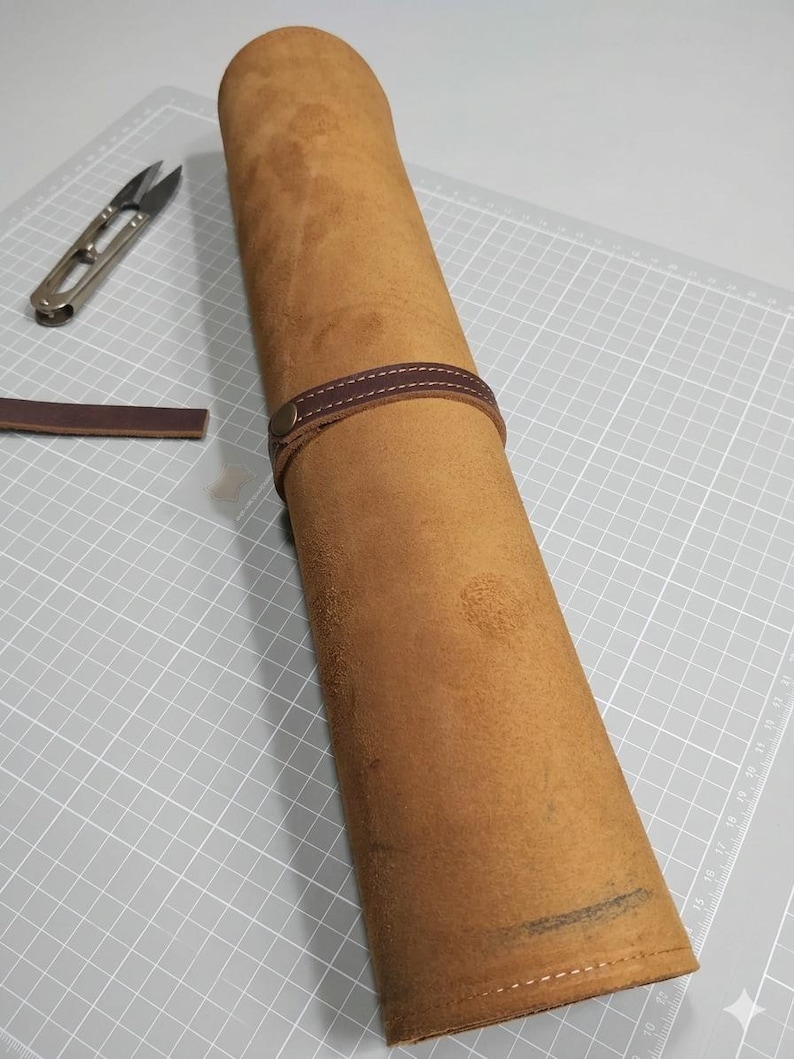 May include: A tan leather roll secured with a dark brown leather strap and a metal snap button. A small pair of metal snips and a loose strip of dark brown leather are positioned nearby on a white cutting mat with a grid pattern.