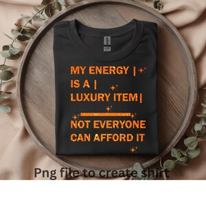 Puede incluir: Camiseta negra con el texto "MY ENERGY IS A LUXURY ITEM NOT EVERYONE CAN AFFORD IT" en letras de purpurina naranja. La camiseta está doblada y colocada sobre una bandeja de madera con hojas decorativas.
