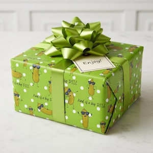 Pode incluir: Uma caixa de presente quadrada embrulhada em papel verde decorada com picles de desenho animado com óculos de sol e bolinhas brancas. Possui uma fita e um laço verdes e uma etiqueta com "Enjoy!"