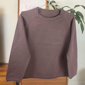 Könnte beinhalten: Ein einfarbiger, mauvefarbener Pullover mit Rundhalsausschnitt und langen Ärmeln. Der Pullover hängt an einem goldenen Kleiderbügel. Der Hintergrund umfasst eine Holzkommode, ein gerahmtes Bild und eine grüne Pflanze.