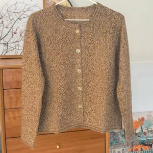 May include: A brown button-down cardigan with a speckled texture. The long-sleeved sweater has a round neckline and is fastened with six round, light-colored buttons. The garment is hanging on a white hanger.