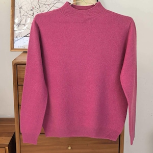 May include: A vibrant pink sweater with a mock neck and long sleeves. The sweater is hanging on a gold hanger. The background includes a wooden cabinet and a framed artwork.