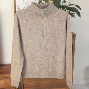 Könnte beinhalten: Ein beigefarbener Pullover mit hohem, geripptem Kragen und einem Viertel-Reißverschluss. Der Pullover hat ein diamantförmiges Strickmuster und lange Ärmel. Das Kleidungsstück hängt an einem Kleiderbügel.