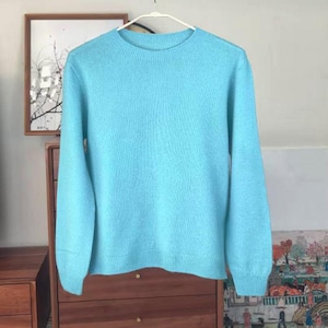 Op de afbeelding: Een lichtblauwe trui met ronde hals en lange mouwen. De trui is gemaakt van een zacht, getextureerd materiaal en hangt aan een witte hanger. De achtergrond omvat een houten kast en ingelijste kunstwerken.