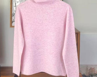 May include: A light pink turtleneck sweater made of a soft, textured material. The sweater is hanging on a hanger. The background includes a wooden cabinet and framed artwork.