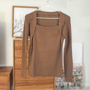May include: A brown ribbed-knit sweater set, comprising a square-neck top and a matching shrug. The set is displayed on a white hanger. The long-sleeved shrug drapes over the top, creating a layered look. The background is neutral.