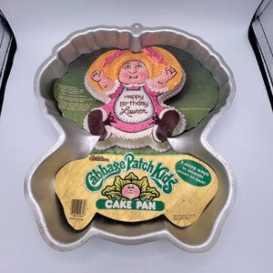 May include: A silver-colored metal Cabbage Patch Kids cake pan by Wilton. The pan includes a cardboard insert featuring a graphic of a decorated doll cake with the text Happy Birthday Lauren, along with the Cabbage Patch Kids logo and branding.