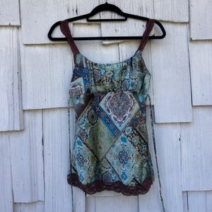 May include: A patterned tank top with a mix of blue, green, and brown hues. The top features a square pattern design with floral and geometric elements. It has brown straps and a brown lace trim at the bottom. The top is hanging on a black hanger.