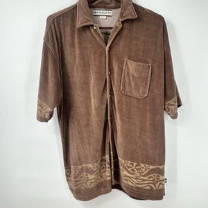May include: A brown velour short-sleeve button-down shirt featuring a collar, a single chest pocket, and a tan patterned band along the hem and sleeve cuffs. The interior neck label displays the brand name Rusty in a white rectangular box.