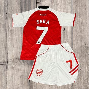 May include: A red and white soccer jersey and shorts set. The jersey has the name "SAKA" and the number "7" on the back. The shorts also feature the number "7". The Arsenal logo is visible on both pieces.
