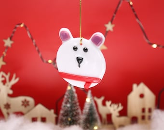 May include: A white polar bear ornament with pink ears and a red scarf hangs against a red backdrop. The bear has black eyes and a black nose. The scene includes miniature houses, trees, and reindeer, suggesting a winter holiday theme.
