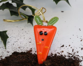 May include: A whimsical orange carrot-shaped ornament with black eyes and a small mouth. The carrot has green leaves and a gold string for hanging. The ornament is set against a backdrop of soil and holly.