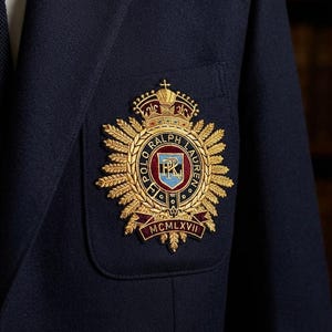 Può includere: Un blazer blu navy con un vistoso stemma ricamato sulla tasca pettorale. L'emblema color oro raffigura una corona, spighe di grano e la scritta Polo Ralph Lauren con uno scudo centrale e un nastro alla base con la dicitura MCMLXVII.