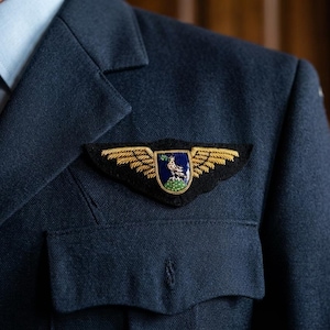 Op de afbeelding: Een donkerblauwe blazer met een lichtblauw overhemd en een donkerblauwe das. Op de borst is een geborduurde badge bevestigd met gouden vleugels die een blauw schild flankeren, voorzien van een witte vogelafbeelding op een groene basis.
