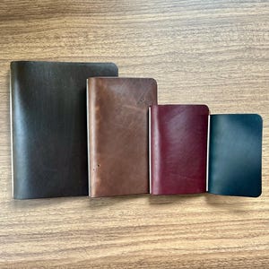 May include: Four rectangular leather notebook covers arranged in descending order of size. The covers are colored dark brown, medium brown, deep burgundy, and dark teal, each featuring a smooth, matte finish and rounded corners.