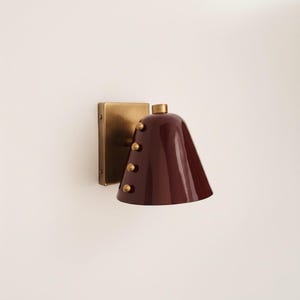 May include: A wall-mounted sconce featuring a square brass backplate and a deep burgundy, bell-shaped shade. The shade is decorated with three small, gold-coloured rivets. The sconce has a gold-coloured top and is designed for indoor lighting.