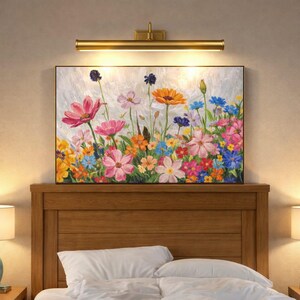 May include: A framed painting of colorful flowers in full bloom. The artwork displays pink, orange, yellow, and blue flowers against a textured white backdrop. A gold-toned picture light is mounted above the painting.
