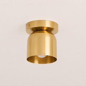 May include: A gold-colored, modern flush mount ceiling light fixture. The fixture has a cylindrical base with a rounded, bell-shaped shade. A small, round light bulb is visible at the bottom. The fixture is mounted on a white ceiling.