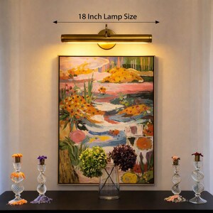 May include: A brass-toned 46 cm picture light mounted above a rectangular painting featuring a colourful abstract landscape. Below the painting, a clear glass vase holds green and purple hydrangeas, flanked by four clear glass candle holders.