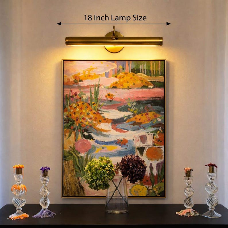 May include: A brass-toned 46 cm picture light mounted above a rectangular painting featuring a colourful abstract landscape. Below the painting, a clear glass vase holds green and purple hydrangeas, flanked by four clear glass candle holders.
