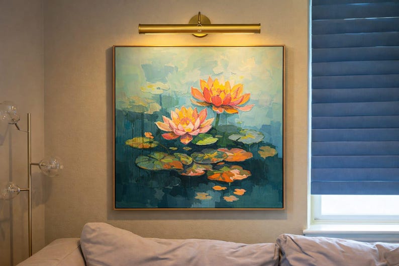 May include: A framed painting shows two orange and pink lotus flowers with lily pads, set against a teal background. A gold picture light shines on the artwork. The painting is above a sofa. The artwork is approximately 60 cm x 60 cm.