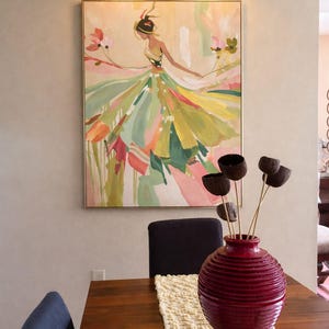 May include: An abstract painting of a woman in a flowing dress, with a gold frame, hangs on a wall. A red vase with dried flowers sits on a wooden table with a cream-coloured runner. A gold light fitting is above the painting.
