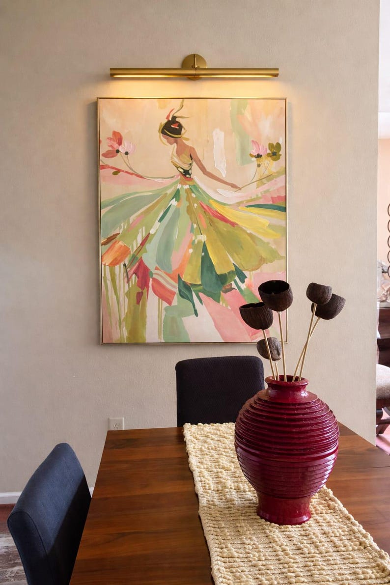 May include: An abstract painting of a woman in a flowing dress, with a gold frame, hangs on a wall. A red vase with dried flowers sits on a wooden table with a cream-coloured runner. A gold light fitting is above the painting.