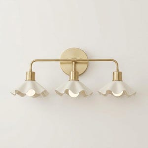 May include: A three-light vanity fixture with a brass finish. The fixture features three white, ruffled-edge shades. The light bulbs are visible within the shades. The fixture is mounted on a round brass backplate.