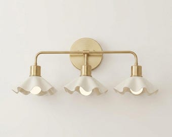 May include: A three-light vanity light fixture with a brass finish. It features three white, ruffled-edge shades. The light bulbs are visible within the shades. The fixture is mounted on a round brass backplate.