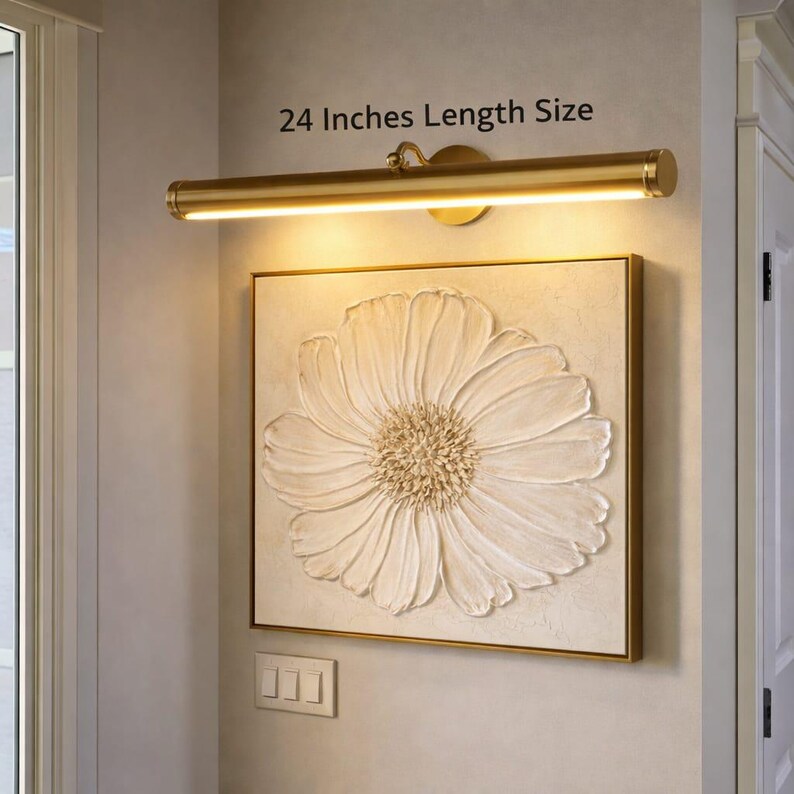 May include: A gold-coloured picture light, 61 cm in length, shines on a framed artwork depicting a large, textured white flower. The artwork is mounted on a neutral wall, with a light switch below.