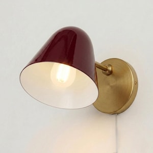May include: A wall-mounted lamp featuring a deep red, cone-shaped shade and a brass-coloured base. The interior of the shade is white, reflecting the light from the exposed bulb. The lamp is designed for indoor use.