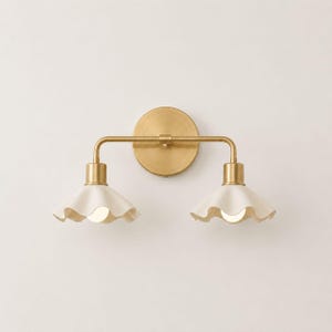 May include: A wall-mounted light fixture featuring a circular brass backplate and a horizontal brass arm supporting two downward-facing, off-white, ruffled ceramic shades. Each shade houses a single light bulb, creating a symmetrical design.