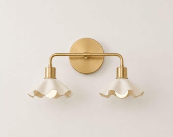 May include: A wall-mounted light fitting featuring a circular brass backplate and a horizontal brass arm supporting two downward-facing, off-white, ruffled ceramic shades. Each shade houses a single light bulb, creating a symmetrical design.