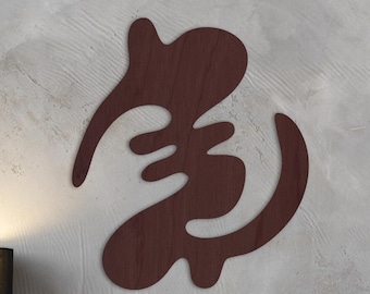 May include: A dark brown wooden Adinkra symbol, mounted on a grey textured wall. The symbol has a unique, flowing design. A black lamp and decorative items are visible below the symbol.